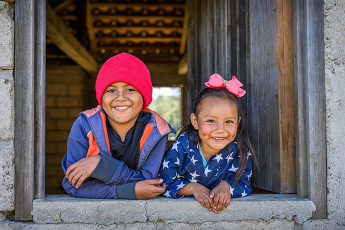 A child in yellow plaid smiles, looking away from the camera, with grey skies and trees in the background.